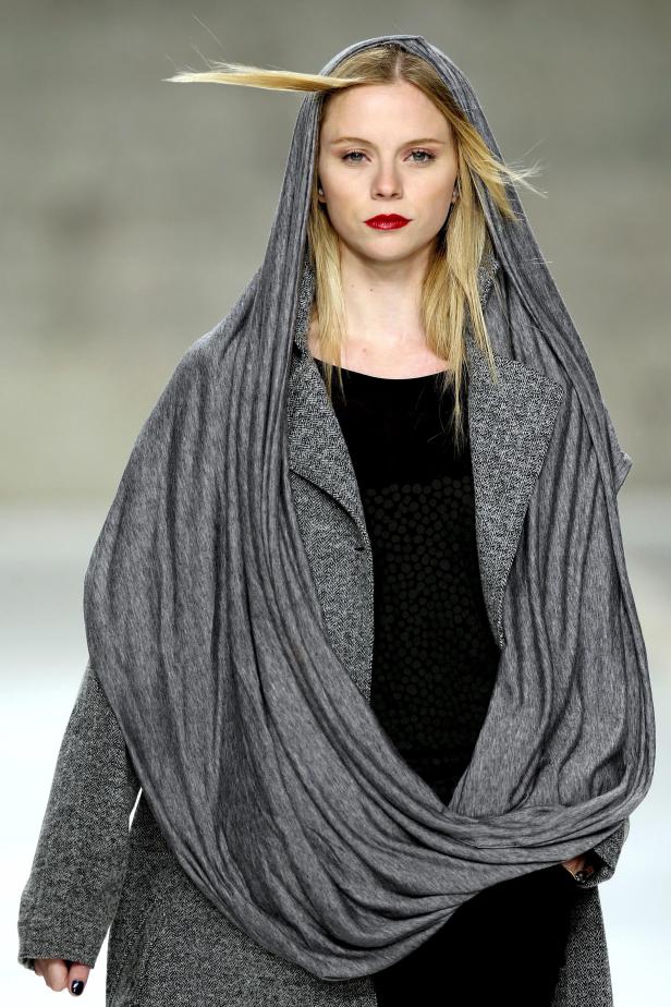 Eine blonde Frau mit rotem Lippenstift trägt einen grauen Kapuzenpullover.