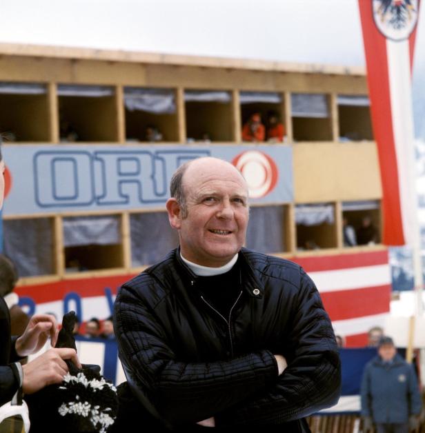 Ein Mann mit verschränkten Armen vor einer Tribüne mit österreichischer Flagge.