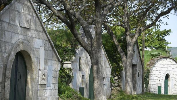 Mehrere traditionelle Weinkeller aus Stein stehen unter Bäumen.