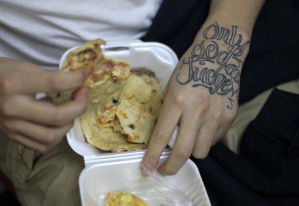 Eine Person isst Pupusas aus einer Styroporbox; die Hand hat ein Tattoo.