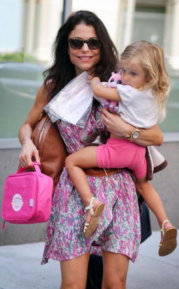 Eine Frau mit Sonnenbrille trägt ein kleines Mädchen und eine pinkfarbene Lunchbox.