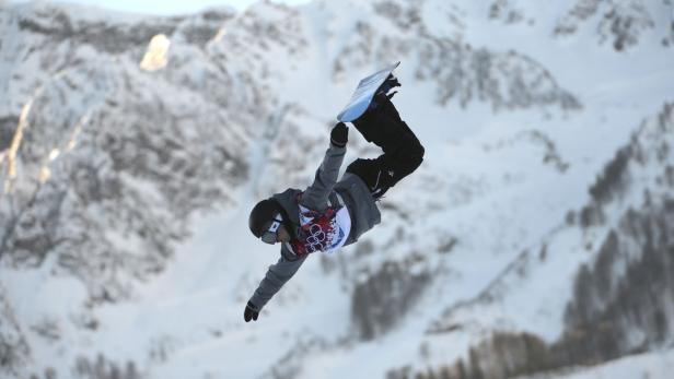 Ein Snowboarder führt vor einer verschneiten Bergkulisse einen Trick in der Luft aus.