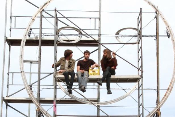 Drei Personen sitzen auf einem Gerüst vor einem großen, kreisförmigen Objekt.