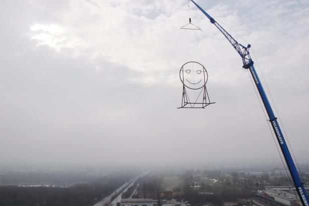 Ein Kran hebt eine große, lächelnde Smiley-Skulptur in der Luft.