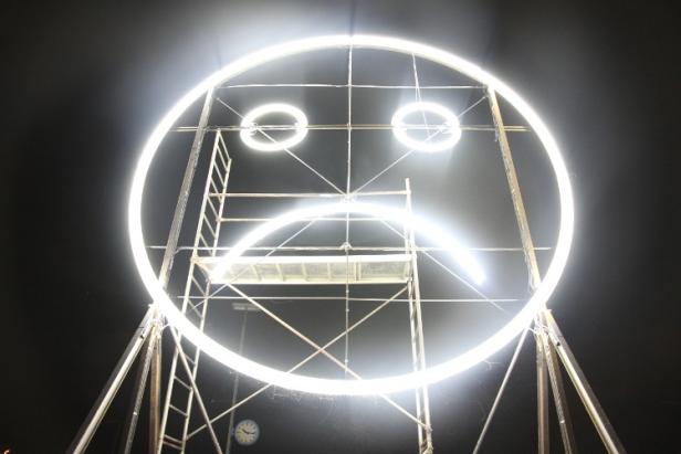 Eine leuchtende, traurige Smiley-Skulptur steht auf einem Gerüst.