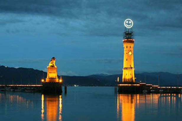 Der Lindauer Leuchtturm mit einem Smiley-Symbol auf der Spitze und der bayerische Löwe am Hafen bei Nacht.
