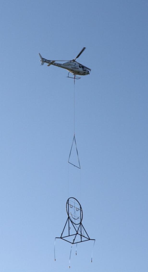 Ein Hubschrauber transportiert eine große, drahtige Skulptur eines Gesichtes mit einem Dreieck darüber.