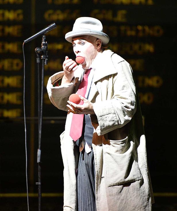 Ein Clown mit weißem Gesicht und Hut jongliert mit roten Bällen vor einem Mikrofon.