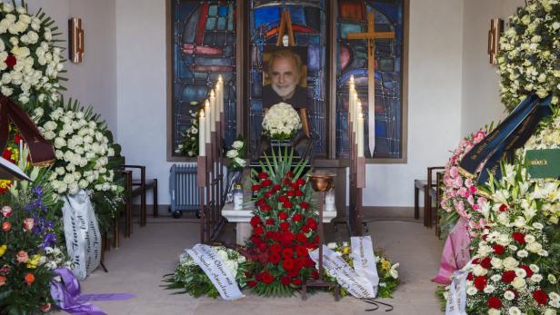 Ein Gedenkaltar mit einem Porträt, Blumenkränzen und Kerzen in einem Raum.