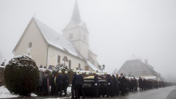 Ein Trauerzug mit Feuerwehrleuten trägt einen Sarg an einer Kirche vorbei im Nebel.