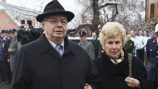 Ein älteres Paar in Mänteln und formeller Kleidung steht vor einer Menschenmenge.