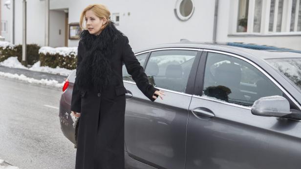 Eine Frau in einem schwarzen Mantel öffnet die Tür eines grauen Autos.