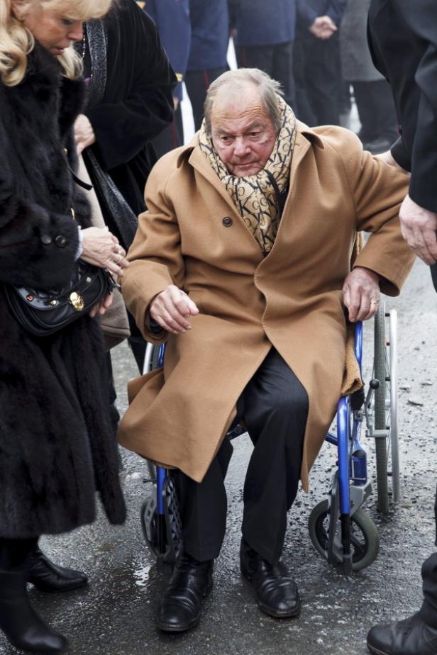 Ein älterer Mann im Rollstuhl, gehüllt in einen beigen Mantel und einen gemusterten Schal.