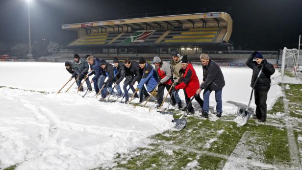 Eine Gruppe von Männern räumt Schnee von einem Fußballfeld vor einem Stadion.