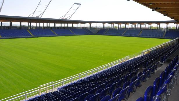 Blick auf ein leeres Fußballstadion mit blauem Gestühl und grüner Rasenfläche.