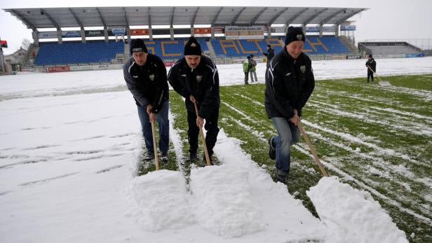 Drei Männer schaufeln Schnee von einem Fußballfeld vor einer Tribüne.