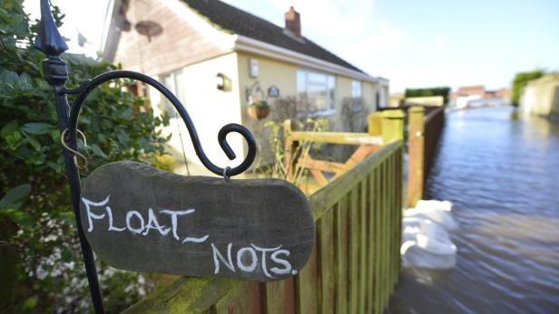 Ein Holzschild mit der Aufschrift „Float Nots“ hängt vor einem überfluteten Haus.
