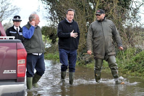David Cameron watet mit anderen Personen durch ein überflutetes Gebiet.
