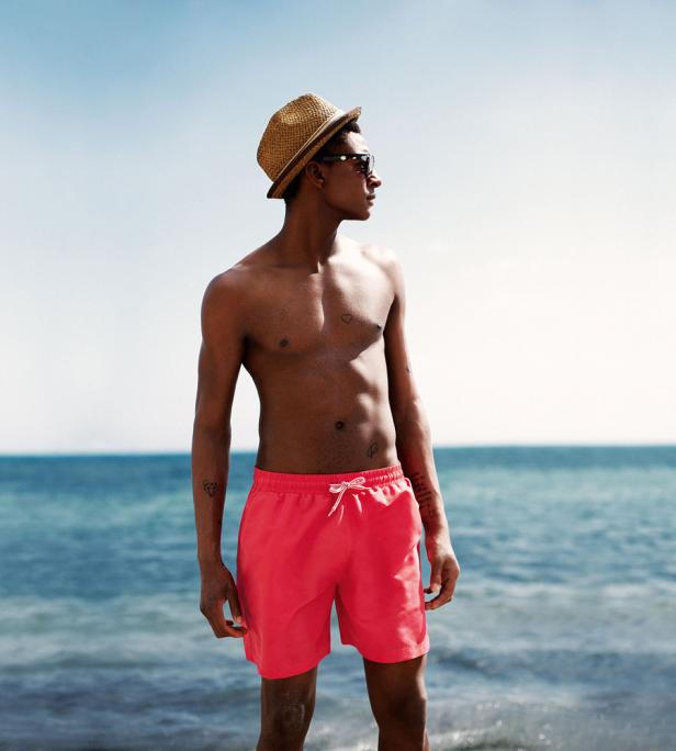 Ein Mann mit Hut und roter Badehose steht am Strand und blickt aufs Meer.