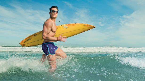 Ein Mann mit Sonnenbrille rennt mit einem Surfbrett ins Meer.