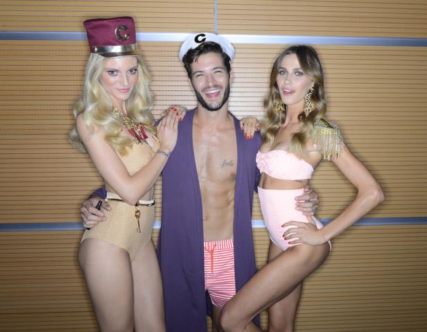 Zwei Frauen und ein Mann posieren in Badebekleidung und auffälligen Accessoires vor einer Wand.