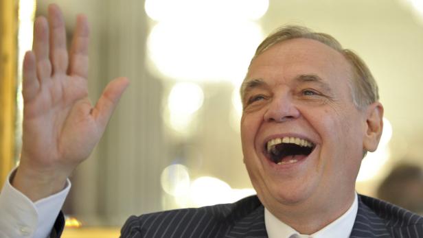 Ein lachender Mann im Anzug hebt die Hand.
