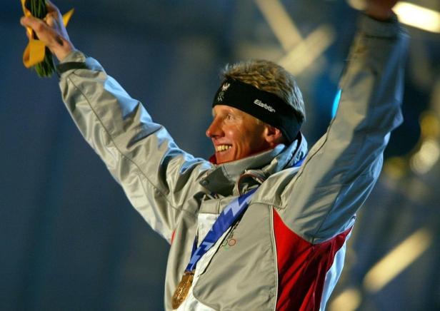 Ein lächelnder Mann mit Goldmedaille und Stirnband reckt die Arme in die Höhe.