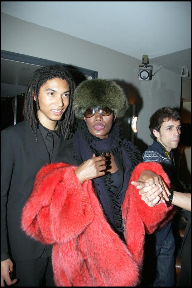 Grace Jones mit rotem Pelzmantel und Pelzmütze, flankiert von zwei Männern.
