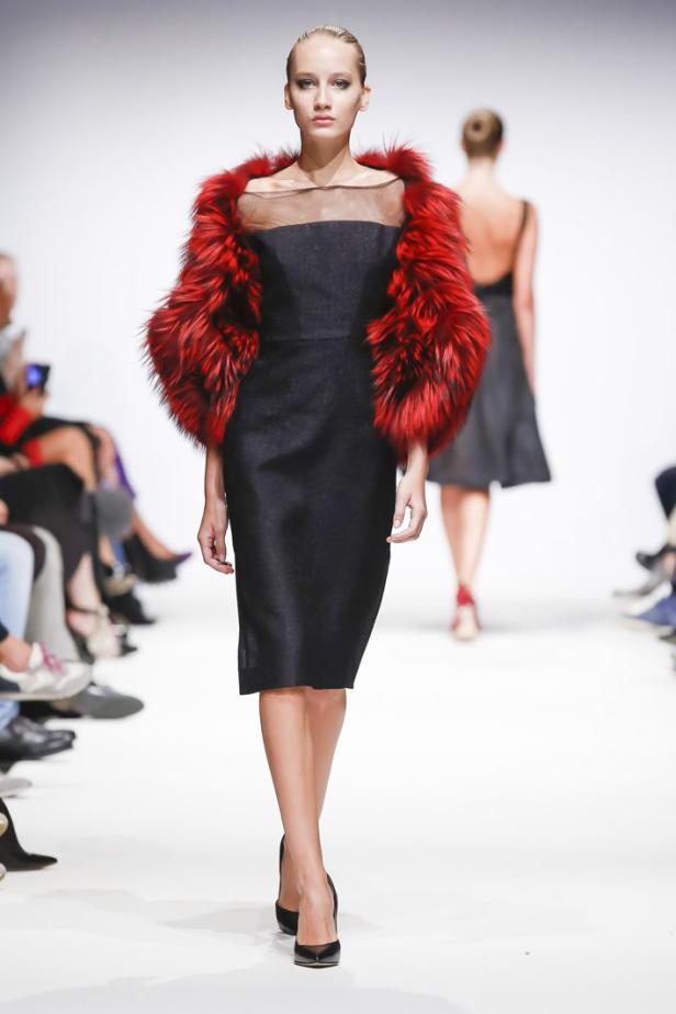 Eine Frau auf dem Laufsteg präsentiert ein schwarzes Kleid mit rotem Pelzbesatz.