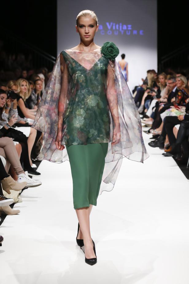 Ein Model präsentiert ein grünes Kleid mit floralem Überwurf auf dem Laufsteg.