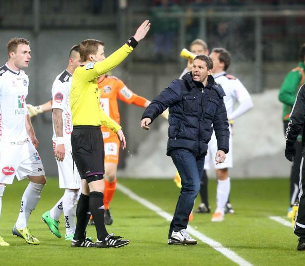 Ein Fußballtrainer gestikuliert auf dem Spielfeld, während ein Schiedsrichter daneben steht.