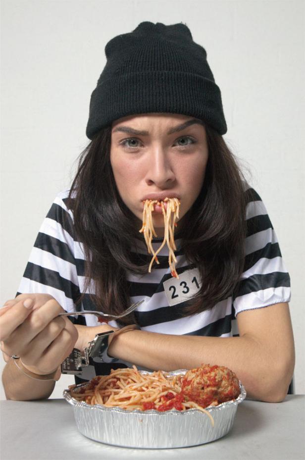 Eine Frau in Sträflingskleidung und Handschellen isst Spaghetti mit einem großen Fleischbällchen.