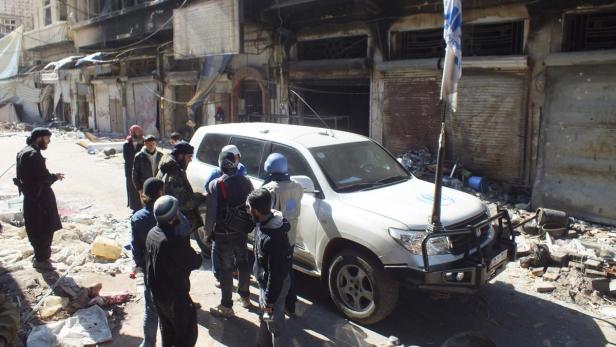 Ein weißes UN-Fahrzeug steht vor zerstörten Gebäuden, umgeben von Menschen.