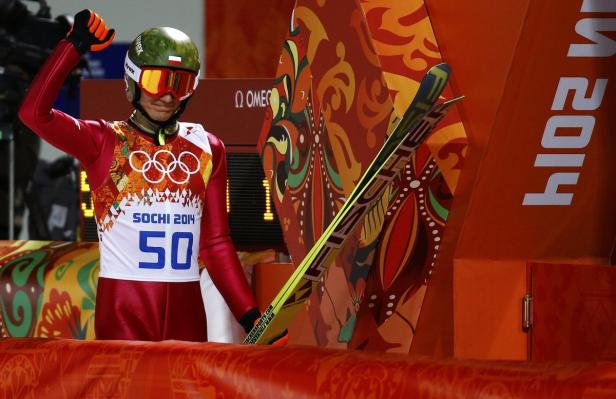 Ein Skispringer im roten Anzug und Helm steht mit erhobenem Arm bereit.