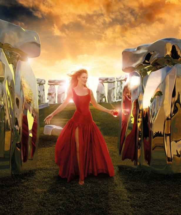 Eine Frau im roten Kleid steht vor einer Installation, die an Stonehenge erinnert, bei Sonnenuntergang.