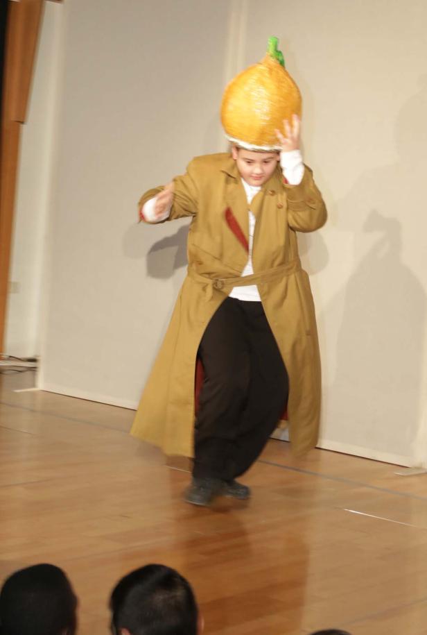 Ein Kind in einem Trenchcoat und einer Zwiebelkopfbedeckung steht auf einer Bühne.