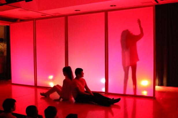 Eine Bühnenaufführung mit Silhouetten vor einem rot beleuchteten Hintergrund.