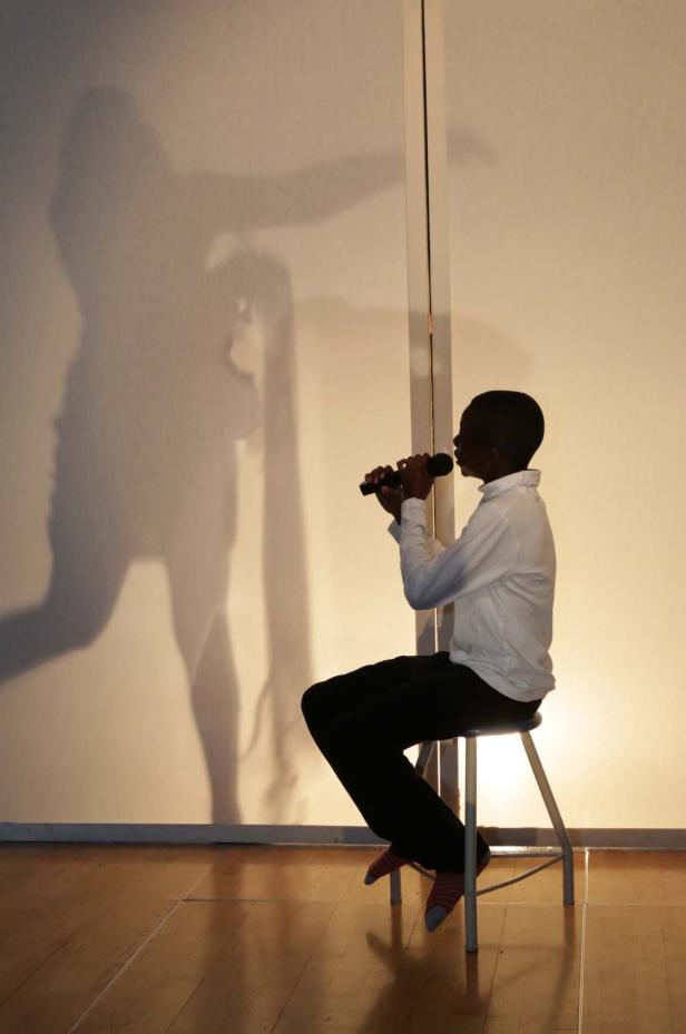 Ein junger Mann sitzt auf einem Hocker und singt in ein Mikrofon, während sein Schatten an die Wand projiziert wird.
