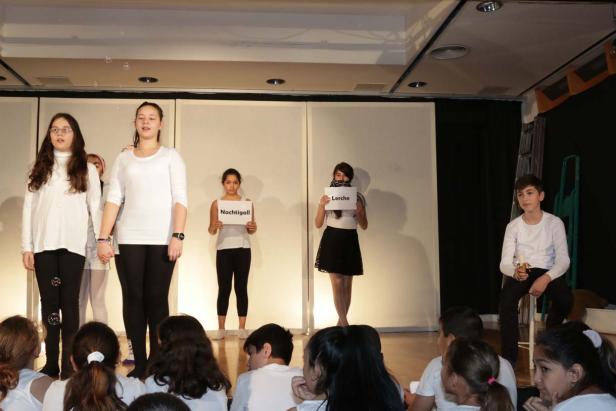 Kinder führen ein Theaterstück auf, einige halten Schilder mit den Aufschriften „Nachtigall“ und „Lerche“ hoch.