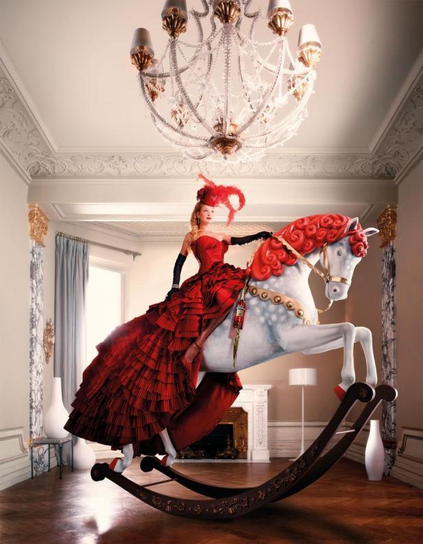 Eine Frau in einem roten Kleid reitet auf einem Schaukelpferd in einem opulenten Raum.