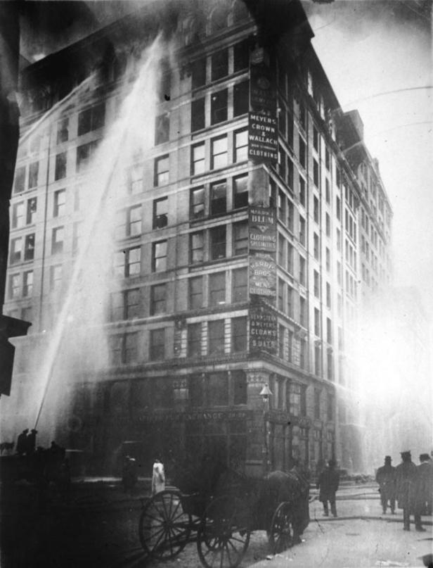 Ein historisches Foto zeigt ein mehrstöckiges Gebäude, das von Feuerwehrleuten mit Wasserwerfern gelöscht wird.