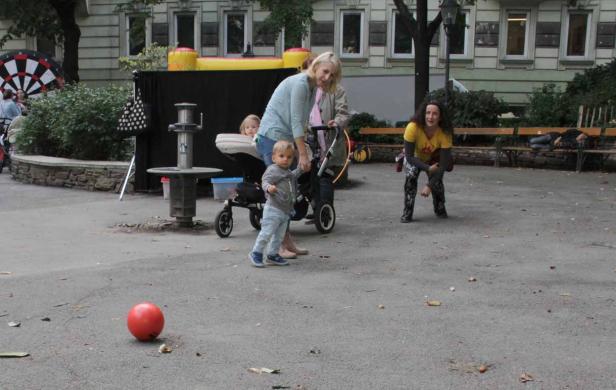 Eine Frau mit Kinderwagen und zwei Kinder in einem Park, im Vordergrund ein roter Ball.