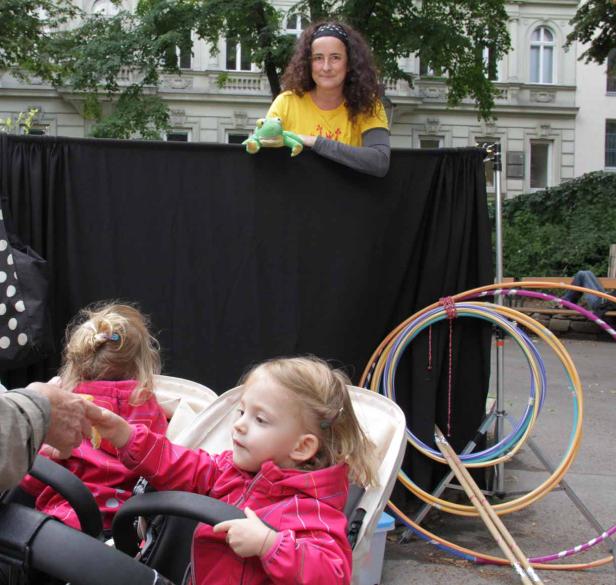 Eine Frau mit einer Froschpuppe steht hinter einer schwarzen Leinwand, während zwei kleine Mädchen in einem Kinderwagen sitzen.