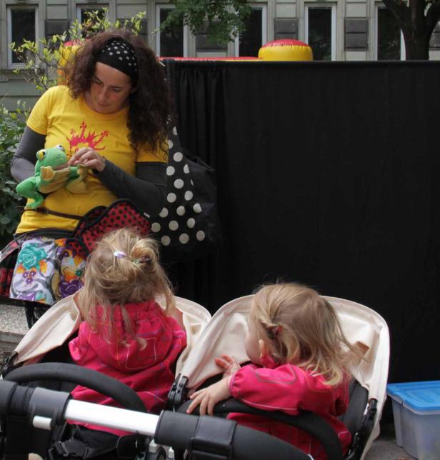 Eine Frau mit einem Stofftier vor einem schwarzen Vorhang, während zwei Kinder in einem Zwillingskinderwagen sitzen.