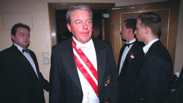 Ein Mann in formeller Kleidung mit einer roten Schärpe steht im Mittelpunkt einer Veranstaltung.