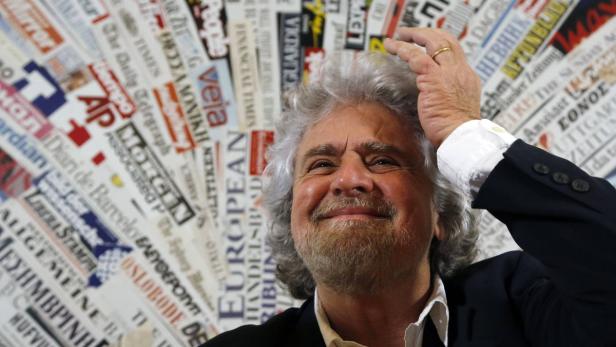 Beppe Grillo lächelt vor einer Wand mit Zeitungsausschnitten.