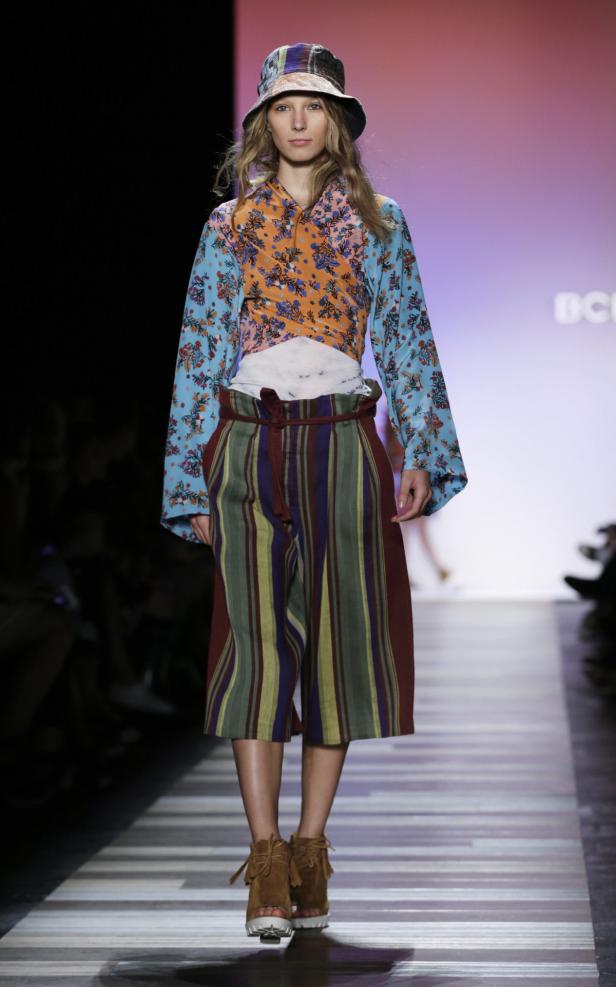 Eine Frau mit Hut präsentiert ein farbenfrohes Outfit auf einem Laufsteg.