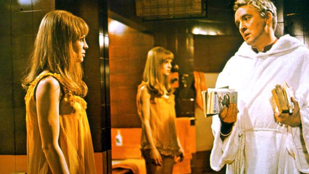 Eine Szene mit zwei jungen Frauen und einem Mann im Bademantel, der Bücher hält.