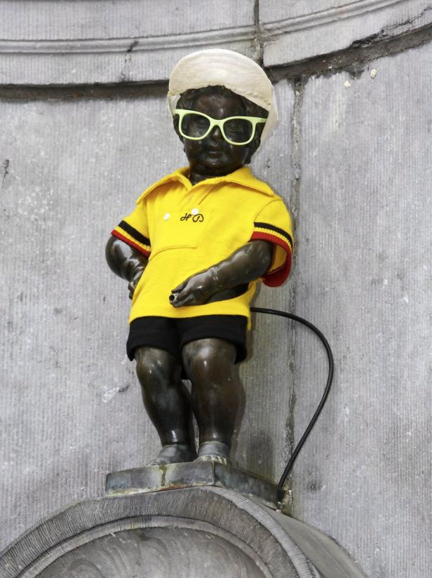 Die Statue Manneken Pis in Brüssel trägt ein gelbes Hemd, eine Mütze und eine Sonnenbrille.