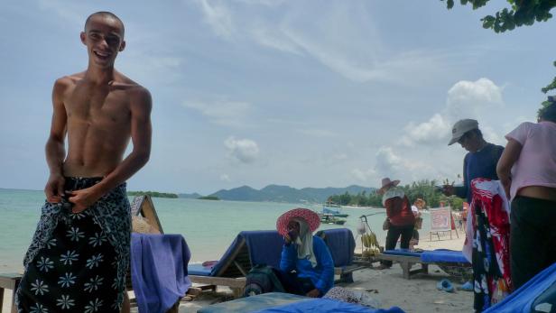 Ein junger Mann mit einem Sarong steht an einem Strand mit Liegestühlen und anderen Personen.
